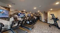This fitness center is designed to offer a comprehensive workout experience within a thoughtfully appointed space. The room is anchored by a striking geometric accent wall clad in a textured, stone-inspired panel design in neutral tones of gray, black, and white, creating a bold visual backdrop for the cardio equipment arranged in front of it. Multiple large flat-screen televisions are mounted along this wall, ensuring entertainment is accessible from every piece of cardio equipment.

The cardio zone features a row of modern treadmills with yellow accent lighting and individual display consoles, accompanied by elliptical trainers and a recumbent bike. Patterned, speckled carpeting runs throughout the space, adding a contemporary aesthetic while providing underfoot comfort.

A dedicated strength training area is situated near floor-to-ceiling windows, which introduce abundant natural light and outdoor views. This zone is equipped with a multi-function cable machine, a Smith machine, a weight bench, and a full dumbbell rack backed by a wall-length mirror. Ceiling fans are distributed throughout, supplementing the recessed lighting and supporting air circulation. Additional amenities include a dual water fountain station and a bank of personal storage lockers, rounding out the functional offerings of this well-designed facility.