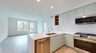 Located at 310 Del Sol Dr in San Diego's Mission Valley neighborhood, this single-floor residence showcases a clean, contemporary design throughout. The open-concept living and kitchen area is anchored by a sleek peninsula with a light wood-accented base and white quartz countertops, complemented by sage-green flat-front cabinetry, open wood shelving, a penny-tile backsplash, and a full suite of stainless steel appliances including a gas range, built-in microwave, dishwasher, and French door refrigerator. Recessed lighting and light hardwood-style flooring extend throughout the living spaces, creating a bright and cohesive atmosphere. Large windows and a sliding glass door in the living area frame views of palm trees and the surrounding streetscape, filling the space with abundant natural light. The bedrooms are well-proportioned with clean white walls and the same warm flooring, offering a calm, neutral palette. One bedroom includes access to a generous walk-in closet with multi-rail storage and a window for natural light. The primary bathroom features large-format stone-look tile flooring, a long wood vanity with a white countertop, and backlit mirrors, while the secondary bathroom includes a full tub-shower combo with white subway tile surround.