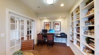 This single-story business center offers a thoughtfully designed workspace that balances functionality with a warm, inviting aesthetic. The room features a built-in wall-mounted desk along the back wall, accommodating two complete computer workstations with monitors, a shared printer, and organized cable management. Two cushioned rolling chairs in a classic red-and-dark-wood finish provide comfortable seating at the workstation.

A floor-to-ceiling built-in bookshelf lines the right wall, offering multiple open shelves stocked with books and decorative items, adding both utility and visual warmth to the space. The walls are adorned with two framed artworks above the desk, contributing a refined, professional atmosphere.

Entry to the room is through elegant French doors with glass panes, allowing visibility into the adjacent space while maintaining a degree of separation. Recessed ceiling lighting and a flush-mount ceiling fixture provide ample, even illumination throughout. Rich, dark carpeting grounds the room, complementing the neutral cream walls and dark wood furnishings. The overall ambiance is polished and professional, well-suited for focused work or study.