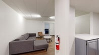 This thoughtfully designed 2-bedroom, 1-bathroom residence spans a single floor and showcases a clean, modern aesthetic throughout. Warm hardwood-style flooring flows consistently across the space, complemented by crisp white walls and panel ceilings fitted with track lighting that provides even, adjustable illumination in every room.

The combined kitchen and living area forms the social heart of the home. The kitchen features gray cabinetry with white countertops and stainless steel appliances, including a full-size refrigerator. The adjoining living room offers a comfortable, open layout with access to the rest of the unit.

The spacious primary bedroom is notably generous in size, accommodating multiple beds, dual dark-framed modular storage units with perforated metal panels and built-in drawers, and a dedicated desk workspace. A sliding door provides access to an outdoor area, allowing natural light to fill the room. The second bedroom mirrors this functional approach with similar storage and study furnishings.

The bathroom is finished with light blue tile accents along the walls, a full-size soaking tub with shower, a vanity with white countertop, gray cabinetry, and a large mirror. A fire extinguisher is visibly mounted near the entry, reflecting attention to safety throughout the unit.