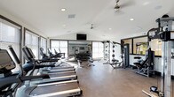 This single-floor fitness center offers a thoughtfully designed workout environment built to support a wide range of exercise routines. The space features soaring vaulted ceilings fitted with ceiling fans, creating an open and airy atmosphere that keeps the room comfortable during intense workouts. Recessed lighting complements the abundant natural light streaming in through large windows fitted with blinds along the perimeter walls.

The cardio zone is prominently featured, housing a row of TRUE-brand treadmills and additional cardio machines arranged for easy access and flow. A dedicated free weights area includes a full dumbbell rack with benches, ideal for strength and toning exercises. On the opposite side, a multi-station cable and weight machine provides options for a comprehensive resistance training workout.

A wall-mounted flat-screen television serves as both an entertainment and informational focal point, positioned above an inspiring typographic mural that adds an energetic, motivational character to the space. The flooring throughout is a durable, dark rubberized surface well-suited for high-traffic gym use. Overall, the fitness center strikes a balance between functionality and modern design, providing a clean, organized, and welcoming environment for users of all fitness levels.