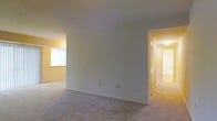 This single-story space offers a straightforward and functional layout designed for comfortable everyday living. The main living area is finished with wall-to-wall carpeting in a neutral tone, providing a soft and cohesive foundation throughout the space. The walls are painted in a warm, light yellow hue that reflects natural light and contributes to a bright, welcoming atmosphere. A window in the living area allows natural light to filter in, enhancing the open feel of the room. A dividing half-wall subtly separates areas within the main living space, adding a sense of structure without sacrificing openness. A hallway visible from the main room leads to additional areas of the space, including what appears to be a closet with louvered doors — a practical storage feature. The hallway is well-lit with overhead lighting, and the carpeting continues seamlessly throughout, maintaining a consistent aesthetic. The overall ambiance is clean, neutral, and move-in ready, with a simple and efficient design that offers a solid canvas for personalization.