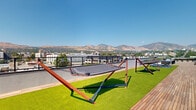 This expansive rooftop terrace is a thoughtfully designed outdoor amenity space that seamlessly blends relaxation, recreation, and natural beauty. The layout is anchored by warm hardwood decking that runs the length of the terrace, complemented by vibrant artificial turf zones that add a lush, green contrast underfoot.

The space is divided into several distinct activity areas. A sun lounge zone features a row of sleek white reclining chaise lounges positioned to capture maximum sunlight, ideal for unwinding outdoors. Nearby, freestanding hammocks with sturdy wooden frames are arranged on the turf, offering a more leisurely way to enjoy the open-air environment. A dedicated seating area with modern Adirondack-style chairs and a central table provides space for social gatherings, while a built-in outdoor kitchen and bar area supports al fresco dining and entertaining.

Generous raised planters filled with ornamental grasses, shrubs, and young trees are distributed throughout the terrace, adding greenery and a sense of privacy. The perimeter is lined with sleek metal railings and wood-panel privacy screens that frame unobstructed, panoramic views of a downtown city skyline in one direction and a dramatic mountain ridgeline in the other. The overall aesthetic is clean, modern, and highly functional, making this rooftop terrace an exceptional outdoor retreat.