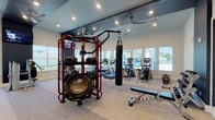 This fitness center is thoughtfully designed to accommodate a wide range of workout styles across two distinct zones. The main exercise area features light-toned carpet flooring, high ceilings with recessed lighting, and ceiling fans that maintain a comfortable atmosphere. Large windows line the walls, drawing in abundant natural light and offering views of the surrounding outdoor landscape. This space is outfitted with a prominent red multi-functional training rig stocked with medicine balls and a suspension trampoline, a hanging heavy bag for boxing training, cable machines, selectorized strength equipment, treadmills, and elliptical trainers. Wall-mounted monitors provide entertainment and instructional content throughout.

A separate, glass-partitioned cycling studio adjoins the main floor, featuring Peloton bikes positioned in front of a large display screen for live and on-demand classes. The studio is finished with light hardwood-style flooring and deep navy accent walls, creating an immersive, boutique-style environment. Natural light filters in through generous windows, including views of palm trees that lend a warm, resort-like feel.

A transitional corridor connects the two zones and includes a water fountain station, restroom access, and additional equipment. The overall design balances modern aesthetics with functional versatility, making it a comprehensive facility for diverse fitness needs.