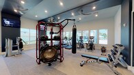 This fitness center is thoughtfully designed to accommodate a wide range of workout styles across two distinct zones. The main exercise area features light-toned carpet flooring, high ceilings with recessed lighting, and ceiling fans that maintain a comfortable atmosphere. Large windows line the walls, drawing in abundant natural light and offering views of the surrounding outdoor landscape. This space is outfitted with a prominent red multi-functional training rig stocked with medicine balls and a suspension trampoline, a hanging heavy bag for boxing training, cable machines, selectorized strength equipment, treadmills, and elliptical trainers. Wall-mounted monitors provide entertainment and instructional content throughout.

A separate, glass-partitioned cycling studio adjoins the main floor, featuring Peloton bikes positioned in front of a large display screen for live and on-demand classes. The studio is finished with light hardwood-style flooring and deep navy accent walls, creating an immersive, boutique-style environment. Natural light filters in through generous windows, including views of palm trees that lend a warm, resort-like feel.

A transitional corridor connects the two zones and includes a water fountain station, restroom access, and additional equipment. The overall design balances modern aesthetics with functional versatility, making it a comprehensive facility for diverse fitness needs.