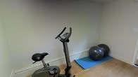 This single-floor fitness space offers a dedicated area for personal exercise and wellness routines. The room features light-toned walls and warm hardwood flooring, creating a clean and welcoming environment for physical activity. An upright stationary exercise bike serves as the centerpiece of the space, providing a reliable option for low-impact cardio workouts. Alongside the bike, two medicine balls and a blue exercise mat are neatly arranged near the baseboard, offering versatility for strength training, core work, and stretching exercises. The neutral wall color and uncluttered layout contribute to an atmosphere that feels calm and focused, minimizing distractions during workouts. The space is efficiently organized to maximize the available floor area, making it well-suited for a variety of fitness routines. The combination of cardio equipment and flexible exercise accessories makes this a functional and practical home gym setup, ideal for individuals seeking a convenient and private space to maintain an active lifestyle.