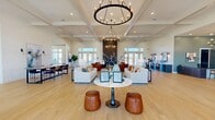 Located in Fort Myers, FL, this single-floor clubhouse at Colonial Gateway Dr offers a generously proportioned, open-plan gathering space designed for both comfort and elegance. The interior is anchored by soaring coffered ceilings adorned with statement ring chandeliers, casting warm light across wide-plank hardwood floors that run throughout the entire space.

The main lounge area features multiple distinct seating arrangements, including plush white sofas, leather armchairs in warm cognac tones, and deep navy swivel chairs, all centered around a dramatic dark-paneled accent wall with a bold floral installation. French doors line the perimeter, flooding the interior with natural light and providing views of the surrounding outdoor areas.

A dedicated bar area showcases a long white marble-topped counter with stylish bar seating, set against a decorative patterned tile backsplash and pendant lighting. Adjacent casual dining areas feature contemporary round tables paired with sculpted wood chairs, ideal for informal gatherings.

A secondary lounge zone, defined by teal accent walls, offers a more intimate setting with a wall-mounted TV and clustered lounge seating. Additional flexible spaces include what appears to be a business or leasing office area with private rooms. The overall ambiance is polished yet welcoming, blending coastal-contemporary design with upscale finishes throughout.