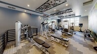 Located at 100 Elena St in Cranston, Rhode Island, this single-level fitness center offers a comprehensive and thoughtfully designed workout facility. The space is organized into distinct training zones, each outfitted to support a variety of exercise routines. The main strength training area features an extensive array of weight machines, cable systems, free weights, and adjustable benches arranged across patterned carpet flooring. A striking decorative ceiling installation, composed of bicycle wheel motifs, adds a creative and energetic design element overhead, complemented by recessed lighting throughout. A dedicated cardio zone houses Matrix-brand treadmills, ellipticals, and stationary bikes positioned in front of large windows that allow natural light and views of the surrounding neighborhood. A separate functional training room features wide-plank wood-style flooring, a full-length wall mirror that amplifies the sense of space, and racks stocked with colorful medicine balls and exercise accessories. A vibrant nature-themed mural adds an inspiring visual backdrop to this area. The facility also includes a welcoming entry corridor with frosted glass panels and tile flooring that leads to an adjoining clubhouse. Throughout the space, bold, large-format murals — including a dynamic cycling-themed graphic — energize the environment and reinforce a motivating athletic atmosphere.