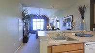 This single-floor residence offers an open-concept design that seamlessly connects the kitchen and living areas, creating a cohesive and inviting atmosphere throughout. The kitchen features warm wood cabinetry, a light-toned countertop, and a stainless steel sink at a peninsula that overlooks the main living space — ideal for both meal preparation and casual entertaining. An elegant chandelier with candelabra-style lighting hangs above, adding a refined decorative touch. The living area is anchored by vaulted ceilings that lend a sense of openness and volume to the space. A ceiling fan provides practical comfort, while large windows dressed with shutters allow an abundance of natural light to fill the room. Neutral carpet flooring and soft wall tones contribute to a calm, welcoming ambiance. Decorative elements such as tall indoor bamboo plants in dark planters, a framed art piece, and tasteful furnishings enhance the overall aesthetic. The combination of functional layout, architectural detail, and thoughtful décor makes this a comfortable and well-designed living space.