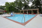 This outdoor amenity space centers around a generously sized covered pavilion situated directly adjacent to a large community swimming pool. The structure features a translucent paneled roof supported by sturdy wooden posts, allowing diffused natural light to fill the space while providing shade and shelter. Dual ceiling fans are mounted overhead, ensuring comfortable airflow during warm weather. The expansive concrete patio floor offers ample open space for movement and gatherings. A bold red modular sectional sofa with wicker-style bases serves as the primary seating feature, positioned along the perimeter wall and providing comfortable lounge space. A low wooden half-wall with decorative iron fencing encloses one side of the pavilion, offering views into a wooded, landscaped buffer area filled with mature trees. Potted evergreen topiaries add greenery and a polished aesthetic throughout the patio. Looking outward from the pavilion, the full expanse of the community pool is visible, complete with poolside lounge chairs, red umbrellas, and well-maintained landscaping. The surrounding residential buildings and tree canopy create a private, resort-like atmosphere, making this covered pavilion an inviting outdoor gathering and relaxation destination.