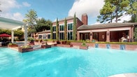 This amenity space combines an expansive outdoor pool area with a thoughtfully designed interior clubhouse. Outside, a large, crystal-clear pool is framed by manicured landscaping, tall cypress trees, and a striking two-story brick building with floor-to-ceiling windows that flood the interior with natural light.

Inside, the clubhouse welcomes visitors with soaring coffered ceilings accented in teal, complemented by warm hardwood-style flooring and sophisticated gray wall paneling. The main lounge area is anchored by a grand stone fireplace with an arched surround, creating a focal point for the generously proportioned seating area furnished with plush chairs and sofas. An elegant chandelier and recessed lighting illuminate the space evenly.

A central granite-topped island with seating serves as a versatile gathering point, while a dedicated coffee and refreshment bar with rich wood cabinetry and granite countertops adds everyday convenience. Wall-mounted televisions provide entertainment throughout the space.

Tall windows along one side of the clubhouse offer direct views of the pool and lush outdoor surroundings, filling seating nooks with natural light. Curated artwork in vibrant blues and teals adds color and visual interest throughout, contributing to the overall refined yet inviting ambiance of this well-appointed community amenity space.