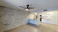 This single-floor space showcases a thoughtfully designed open-concept layout that seamlessly connects the living area to the kitchen. A prominent whitewashed exposed brick accent wall serves as the focal point of the living space, adding texture and character to the otherwise clean, modern aesthetic. Light wood-style flooring runs throughout, complementing the neutral color palette and contributing to a bright, airy atmosphere.

A modern ceiling fan with integrated lighting is centrally mounted on the textured ceiling, providing both functionality and a contemporary design element. The open pass-through kitchen features white cabinetry, light-toned countertops, and a full suite of stainless steel appliances, including a refrigerator, microwave, and range. The kitchen's peninsula-style layout offers additional counter space and provides a natural visual separation between the cooking and living areas. Recessed overhead lighting in the kitchen ensures the workspace is well-lit. HVAC vents are integrated into the ceiling, supporting efficient climate control throughout the space.