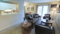 This single-floor residence features an open-concept design that seamlessly connects the living, dining, and kitchen areas, creating a cohesive and functional layout. The living room serves as the centerpiece of the space, anchored by a plush, dark upholstered sofa accented with decorative throw pillows. A round wooden coffee table adds warmth and character to the seating area, complemented by neutral-toned carpeting throughout. A large sliding glass door or wide window floods the living area with abundant natural light while providing views of the outdoors. Framed wall art adds a decorative touch above the dining area, which features a classic wooden table and chairs positioned adjacent to the living room. A ceiling fan with a light fixture overhead ensures comfortable air circulation. The kitchen is partially visible through a pass-through opening, hinting at a practical and connected layout that encourages interaction between spaces. The overall color palette is warm and neutral, with beige walls and dark furnishings creating a balanced, inviting ambiance throughout the home.