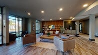 This expansive single-floor amenity space at Barlow offers a thoughtfully designed communal environment that blends comfort, functionality, and modern aesthetics. The welcoming lounge area anchors the space with rich leather sofas, a warm wood-paneled feature wall lined with open shelving, a mounted television, and wood-look flooring underfoot. Adjacent to the lounge, a dedicated coffee bar with cabinetry and countertop space provides a convenient spot for residents to recharge.

The space transitions seamlessly into an entertainment zone featuring a full-size pool table and a separate game room equipped with a poker table, both bathed in natural light from tall windows overlooking lush palm trees and a resort-style pool. Floor-to-ceiling black-framed glass walls open onto the pool deck, flooding the interior with sunlight and creating a seamless indoor-outdoor connection.

A business center area provides workstations with desktop computers set against a textured accent wall with custom artwork, ideal for productivity. A bold teal-accented bar counter with geometric patterned tile flooring adds a vibrant design moment near the main corridor. The entry lobby features comfortable seating arrangements and large windows, creating a bright and inviting arrival experience that ties the entire amenity floor together.