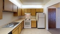 This single-floor residence offers a practical and comfortable layout designed for everyday living. The kitchen is efficiently arranged with warm oak cabinetry providing ample storage, light-toned countertops, tile flooring, and a full suite of appliances including a freestanding electric range, refrigerator, and stainless steel sink — all beneath a well-lit overhead soffit. The living area is generously sized, finished with neutral carpet and bright white walls that create a clean, open atmosphere. Large vertical blinds frame a sliding glass door that opens to an outdoor balcony, allowing natural light to fill the space. The bedrooms are carpeted and offer a quiet, understated feel, with windows fitted with vertical blinds. The primary bedroom includes direct access to a walk-in closet and an en-suite bathroom. Both bathrooms are finished with light tile flooring, wood-toned vanity cabinetry, and walk-in showers enclosed by glass doors. The overall design is straightforward and well-maintained, with a neutral color palette throughout that lends a clean, move-in-ready appeal to the entire space.