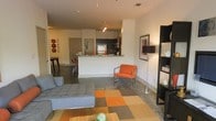 This contemporary single-floor space is defined by its open-concept design, seamlessly connecting the living, dining, and kitchen areas into one cohesive and functional layout. Polished concrete floors run throughout, lending an industrial-modern aesthetic that anchors the space with a clean, sleek foundation.

The living area is generously proportioned, centered around a tufted gray sectional sofa and accented by a bold, multi-toned area rug in shades of orange, tan, and brown. A vibrant orange accent chair adds a lively pop of color, complemented by a dark bookshelf unit along the wall that provides both storage and display space.

The kitchen is positioned at the rear of the open floor plan and features dark cabinetry, stainless steel appliances including a full-size refrigerator, and a breakfast bar with light-toned countertops that doubles as a casual dining surface. Track lighting overhead ensures the kitchen is well-illuminated.

Adjacent to the kitchen, a dining area with a dark wood table is visible, offering a dedicated space for meals. The walls are kept in a clean, neutral white throughout, providing a bright backdrop that highlights the colorful furnishings and wall art. The overall ambiance is modern, urban, and energetic.
