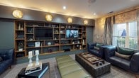 This thoughtfully designed amenity lounge is a single-level space that blends entertainment and relaxation in a refined, upscale setting. The space is divided into distinct yet cohesive zones, each offering a unique experience for residents or guests.

Upon entry, a reception-style anteroom greets visitors with striking metallic textured wall panels, soft carpeted flooring, and a built-in wet bar complete with a beverage cooler and marble-finished countertop. Navy blue leather lounge chairs accented with teal throw pillows provide a stylish and welcoming seating area adjacent to glass-paned French doors.

Through the French doors, the main lounge opens into a generously proportioned entertainment space. A full billiards table with a natural wood base commands the center of the room beneath a contemporary chandelier, while draping curtains frame a glass door that leads to an outdoor area, allowing natural light to filter in. High-top bistro tables with green upholstered chairs line the wall, which features elegant raised panel molding details.

A dedicated media and lounge area is anchored by a floor-to-ceiling built-in shelving wall in warm wood tones, housing dual flat-screen televisions, curated books, and decorative objects. Ambient globe sconces add warm accent lighting, while a backgammon coffee table and plush seating create an inviting atmosphere perfect for social gatherings or quiet relaxation.
