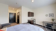 Located at 916 N College Ave in Bloomington, Indiana, this efficiently designed unit offers a thoughtful layout that maximizes every inch of its compact footprint. The main living area serves as both a sleeping and lounge space, featuring a lofted bed with built-in storage drawers underneath, a gray loveseat sofa, and a window with blinds that allows natural light to filter in along with views of the surrounding neighborhood. A wall-mounted HVAC unit provides in-room climate control.

A built-in kitchenette area includes dark espresso-toned upper and lower cabinetry, a granite-style countertop with a sink, and a mini-refrigerator — offering practical in-room convenience. A metal desk and chair provide a dedicated study or workspace. The entryway features a closet and a bold dark-framed front door, contributing to the modern aesthetic of the unit.

The private bathroom is accessible directly from the main room and features the same dark cabinetry with a granite-style vanity top, an undermount sink, a large mirror, a toilet, and a separate shower area. Light-toned tile flooring and crisp white walls give the bathroom a clean, contemporary feel. The overall design is modern and utilitarian, well-suited for student housing near Indiana University.