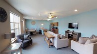 This single-floor space features an open-concept layout that seamlessly integrates a living area, dining space, and kitchenette into one cohesive environment. The walls are painted in a soft, calming blue tone that pairs with neutral gray carpeting to create a relaxed and welcoming ambiance throughout.

The living area is furnished with a comfortable neutral-toned sofa and armchairs arranged around a wooden coffee table, with a large flat-screen television mounted on a media console positioned along one wall. A decorative wall medallion adds a subtle artistic touch to the space.

Adjacent to the living area, the dining section accommodates a dark-toned rectangular table with multiple chairs, lit overhead by pendant lighting that adds a warm, functional glow. A kitchen island with bar seating serves as a transitional element between the dining and kitchenette zones.

The kitchenette is equipped with wood-toned cabinetry, a stainless steel refrigerator, and countertop appliances, making it a practical amenity within the open layout. Recessed lighting and a ceiling fan with an integrated light fixture provide ample illumination and ventilation throughout the space.