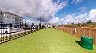 This amenity space combines an outdoor pet park with a comprehensive indoor fitness center, offering a well-rounded recreational experience. The pet park features a generously sized, lush artificial turf area enclosed by a combination of black iron and wooden privacy fencing. Green agility obstacles are positioned throughout the run, providing enrichment for pets. The open layout offers ample room for animals to exercise freely under open skies. The fitness center is a thoroughly equipped, well-lit space bathed in natural light from large windows with views of the surrounding community. The flooring features a distinctive herringbone-patterned carpet tile in blue and grey tones throughout. Ceiling fans and recessed lighting complement the bright interior. The gym is divided into functional zones: a cardio area hosts multiple treadmills and a wall-mounted television, while the strength training area includes a power rack, cable machine, rowing machines, a Precor stationary bike, and a full dumbbell rack. Additional equipment includes medicine balls in multiple weights, battle ropes, and stability balls. Full-length wall mirrors expand the sense of space and allow for proper form checks. The facility is thoughtfully designed to support a wide range of fitness routines in a spacious, organized environment.