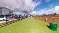 This amenity space combines an outdoor pet park with a comprehensive indoor fitness center, offering a well-rounded recreational experience. The pet park features a generously sized, lush artificial turf area enclosed by a combination of black iron and wooden privacy fencing. Green agility obstacles are positioned throughout the run, providing enrichment for pets. The open layout offers ample room for animals to exercise freely under open skies. The fitness center is a thoroughly equipped, well-lit space bathed in natural light from large windows with views of the surrounding community. The flooring features a distinctive herringbone-patterned carpet tile in blue and grey tones throughout. Ceiling fans and recessed lighting complement the bright interior. The gym is divided into functional zones: a cardio area hosts multiple treadmills and a wall-mounted television, while the strength training area includes a power rack, cable machine, rowing machines, a Precor stationary bike, and a full dumbbell rack. Additional equipment includes medicine balls in multiple weights, battle ropes, and stability balls. Full-length wall mirrors expand the sense of space and allow for proper form checks. The facility is thoughtfully designed to support a wide range of fitness routines in a spacious, organized environment.