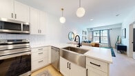 This single-floor residence presents a thoughtfully designed open-concept layout that seamlessly connects the kitchen and living areas. The kitchen is a standout feature, outfitted with white shaker cabinetry, matte black hardware, and bright white quartz countertops. A stainless steel farmhouse sink anchors the peninsula, complemented by a matching stainless steel appliance suite including a refrigerator, range, over-the-range microwave, and dishwasher. A classic white subway tile backsplash and two globe pendant lights with brass accents complete the contemporary aesthetic. Light hardwood flooring flows throughout the kitchen and living areas.

The living room benefits from a large picture window that invites natural light and frames outdoor views, creating an airy and welcoming atmosphere. Two well-appointed bedrooms feature plush carpeting, large windows with outdoor views, and ceiling fans or ample overhead lighting. The bathroom is finished with white shaker cabinetry, a single undermount sink with matte black fixtures, a large vanity mirror, and a shower-tub combination. The consistent use of matte black hardware, crisp white walls, and warm wood tones throughout the space creates a cohesive modern aesthetic.