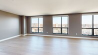 Located at 1 Uptown Cir in Normal, IL, this thoughtfully designed 4-bedroom, 2-bathroom residence offers an impressive single-floor layout with generous square footage throughout. The open-concept kitchen and living room serve as the heart of the home, with the kitchen showcasing rich, dark wood cabinetry extending floor to ceiling, sleek white countertops, and modern hardware — providing abundant storage and workspace. The living area is bathed in natural light through large, floor-to-ceiling windows that frame elevated views of the surrounding cityscape. Light hardwood flooring flows through the main living spaces, complementing the contemporary grey and neutral color palette. The four bedrooms are carpeted for comfort, with the spacious primary bedroom featuring generous closet storage. Additional bedrooms offer natural light and their own closet solutions. Two well-appointed bathrooms include a walk-in glass-enclosed shower with decorative tile accents and clean modern fixtures. A walk-in closet with customizable shelving and hanging systems adds practical storage. A standout feature of this residence is the expansive rooftop terrace, finished with large-format pavers and cable railings, offering sweeping views of the Normal/Bloomington skyline — an exceptional outdoor retreat above the city.