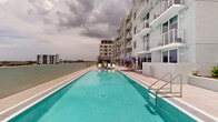 Located at 1025 E 25th Street in Hialeah, Florida, this outdoor amenity space is part of a contemporary multi-story residential building characterized by a clean, modern architectural aesthetic with a neutral grey and white exterior, private balconies, and large windows throughout. The centerpiece of the amenity level is a generously sized swimming pool with crystal-clear turquoise water, stainless steel entry rails, and a surrounding pool deck finished in large-format light-toned porcelain tile. The sun terrace is well-appointed with rows of white chaise lounges shaded by navy blue market umbrellas, offering a resort-like atmosphere ideal for relaxation. Panoramic views of the surrounding Hialeah skyline and open sky are visible from the pool deck, lending an open and airy feel to the outdoor environment. Adjacent to the pool area, a thoughtfully designed courtyard lounge provides a more intimate setting, featuring stylish upholstered seating arrangements set against raised planter beds filled with ornamental grasses and tropical greenery. Sliding glass doors connect the outdoor terrace directly to interior common areas, ensuring seamless indoor-outdoor flow. The overall ambiance is polished and inviting, combining functional amenities with landscaped natural elements in a well-maintained, modern South Florida setting.