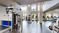 This expansive fitness center is designed to support a full range of workout routines in a bright, contemporary setting. The space features soaring double-height ceilings that lend an open, airy feel, complemented by large floor-to-ceiling windows and elevated clerestory windows that flood the interior with abundant natural light and offer views of the surrounding streetscape and greenery.

The cardio zone is well-appointed with multiple treadmills, elliptical trainers, stair climbers, a rowing machine, and a stationary air bike, all positioned to take advantage of the panoramic window views. A dedicated functional training area includes a TRX suspension training rig, providing space for bodyweight and suspension-based exercises.

The strength training section features a comprehensive selection of selectorized cable machines, plate-loaded equipment, and a full dumbbell rack spanning a wide range of weights, accompanied by large wall mirrors that enhance the sense of space. Recessed and linear LED lighting illuminates the strength area with a modern, focused ambiance.

The interior design balances functionality with aesthetic appeal, showcasing a motivational typography wall graphic, neutral tones, durable rubber and hard-surface flooring, and structural columns that define distinct workout zones. The overall atmosphere is clean, energizing, and purpose-built for fitness.