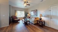 This single-story residence offers a thoughtfully designed layout that balances comfort and functionality across its key living spaces. The living room serves as a central gathering area, featuring rich hardwood flooring, a ceiling fan, and large windows with vertical blinds that allow generous natural light to filter in. An adjacent dining area connects seamlessly to the kitchen, centered beneath a pendant light and positioned next to a sliding glass door for additional brightness.

The galley-style kitchen is efficiently organized with warm wood cabinetry extending to the ceiling, grey countertops, a stainless steel double sink, and a full suite of black and stainless steel appliances including a range, microwave, refrigerator, and dishwasher. A bamboo-shaded window adds a natural accent.

The bedrooms are carpeted for comfort, each equipped with a ceiling fan and ample space. One bedroom includes a walk-in closet with built-in shelving. Both bathrooms feature hardwood-style flooring, wood-toned vanities with wide mirrors, and a tub-shower combination with tile surround. Throughout, the home maintains a warm, neutral palette with consistent finishes that create a cohesive and inviting atmosphere.