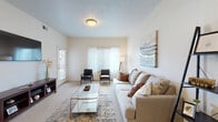 This single-floor residence offers a thoughtfully designed layout with a cohesive, neutral color palette and carpet flooring throughout the main living and bedroom areas. The spacious living room serves as the central gathering space, featuring a textured ceiling, wall-mounted flat-screen television, and multiple windows dressed with sheer curtains and horizontal blinds that allow ample natural light. Two mid-century-style armchairs complement a large sectional sofa arranged around a glass-top coffee table, creating a comfortable and functional seating area.

The kitchen is well-equipped with light maple cabinetry, dark countertops, and black appliances including a range and over-the-range microwave. A peninsula with bar seating provides additional dining space and opens toward a connected dining or utility area. Wood-look flooring anchors the kitchen zone.

The bedrooms are carpeted and bright, each benefiting from natural light through sizable windows. One room features a dedicated workspace with a desk, while another is furnished with a daybed, offering flexibility of use.

Two full bathrooms include single-sink vanities with light wood cabinetry, large mirrors, tiled flooring in a stone-look finish, and shower-tub combinations, providing practical and clean functional spaces throughout.