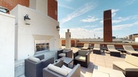 This rooftop terrace offers a well-appointed outdoor retreat elevated above a vibrant urban landscape. The space is designed to accommodate both relaxed lounging and social gatherings, with distinct zones thoughtfully arranged across a large, light-toned tile floor. A striking built-in outdoor fireplace anchors the main seating area, framed by white stucco walls and flanked by warm brick facades. Around it, comfortable wicker armchairs and sofas with cream cushions create an inviting conversation area. A generous row of padded sun loungers lines one side of the terrace, ideal for taking in the open sky and city views that extend in multiple directions, including panoramic sightlines of a downtown skyline with high-rise towers. Concrete pillar accents and low brick perimeter walls contribute to the architectural character of the space. Wall-mounted lantern-style light fixtures suggest the terrace is equally functional after sundown. The overall ambiance balances urban energy with relaxed outdoor comfort, making it a versatile amenity space suited for sunbathing, evening gatherings, or simply enjoying the open-air setting above the city.