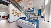 This generously scaled amenity space is defined by its dramatic double-height ceilings and open, airy layout that spans a single floor. The entrance foyer welcomes visitors with floor-to-ceiling glass doors, oversized framed artwork, and lush indoor greenery, establishing a refined yet approachable aesthetic from the outset.

The central gathering area commands attention with a striking large-scale mixed-media mural in deep blue and white tones anchoring the back wall. Oversized drum pendant lights hang at varying heights above the polished large-format tile flooring, casting warm, even light across the space. A stylish bar with barstool seating extends along this focal wall, flanked by warm wood cabinetry.

A dedicated lounge zone features contemporary sectional sofas, bold cobalt accent chairs, and a plush area rug, creating an inviting setting for socializing. A billiards table serves as a centerpiece for the recreation area, complemented by a wall-mounted television nearby.

A sculptural staircase with warm wood accents and black metal railings leads to a mezzanine overlook, adding vertical drama. A secondary lounge benefits from tall windows that flood the area with natural light and offer outdoor views. Throughout, curated artwork, statement planters, and cohesive blue-toned accents unify the space with a contemporary, design-forward character.