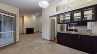 This single-floor residence showcases a thoughtfully designed open layout with a mix of carpet and hardwood-style flooring throughout. The living area is a standout space, featuring soaring vaulted ceilings, multiple windows with views of lush surrounding trees, and a classic wood-framed fireplace that anchors the room. Baseboard heating lines the walls, and natural light flows freely through the generously sized windows fitted with horizontal blinds.

The kitchen is well-appointed with dark espresso cabinetry, granite-style countertops, stainless steel appliances including a refrigerator and range, and a double-basin sink positioned beneath a window. Glass-front upper cabinets add an elegant touch, and pendant lighting completes the functional yet stylish space.

A pass-through counter with a decorative ledge connects the kitchen area to the living space, enhancing the open-concept feel. The bathroom features a dark vanity with a wide countertop, a large mirror, and hardwood-style flooring. A dedicated laundry area houses a full-size washer and dryer behind bifold doors. Additional rooms include a carpeted bedroom with direct access to a bathroom, rounding out this well-organized and comfortable residence.