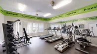 This fitness center is a well-appointed, single-room exercise facility designed to accommodate a full range of workout activities within a compact footprint. The space is thoughtfully organized to maximize functionality, featuring a dedicated cardio zone with multiple treadmills and an elliptical machine, as well as a strength training area equipped with a multi-station cable machine, a weight bench, and a tiered dumbbell rack offering a variety of free weight options. A large wall-to-wall mirror along one side of the room enhances the sense of space and allows users to monitor their form during exercise. Natural light filters in through slatted window blinds, complementing the overhead fluorescent lighting and ceiling fans that keep the environment comfortable. The flooring consists of durable, dark rubberized material suited for high-traffic gym use. A standout design feature is the bold green motivational banner that wraps around the perimeter of the room near the ceiling, displaying energizing words and phrases such as "Determination," "Commitment," "Perseverance," and "Improve." The textured ceiling and clean white walls provide a neutral backdrop that keeps the focus on the equipment and the uplifting décor. Posted weight room rules and a hand sanitizer dispenser reflect a commitment to cleanliness and safety.
