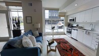 This single-floor unit showcases a thoughtfully designed open-concept layout that efficiently combines the living, dining, and kitchen areas into one cohesive space. Light hardwood-style flooring runs throughout, lending a clean, contemporary aesthetic that ties the zones together visually.

The living area features a teal sofa accented with decorative throw pillows, a round wood-and-metal coffee table, and a vibrant patterned area rug that adds warmth and character to the otherwise neutral palette. A floor lamp and framed wall art contribute a curated, residential feel.

At the center of the space, a floor-to-ceiling window allows an abundance of natural light to pour in, offering street-facing views of the surrounding urban environment. A compact round dining table with modern wire-frame chairs is positioned directly in front of this window, maximizing both light and ambiance.

The kitchen occupies the adjacent area and is outfitted with white cabinetry, stainless steel appliances including a range and over-the-range microwave, and ample counter space. The overall layout is highly functional, making efficient use of the available footprint while maintaining an open, airy atmosphere throughout.