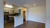 This single-floor space showcases a thoughtfully designed open-concept layout that seamlessly connects the kitchen and living area. The kitchen is outfitted with light wood cabinetry, a dark subway tile backsplash, and a light-toned countertop peninsula that doubles as a breakfast bar, providing both additional prep space and a casual dining option. A stainless steel sink is integrated into the peninsula, adding to the kitchen's functional design. Recessed lighting illuminates the space, complementing the neutral, light grey walls throughout.

Dark hardwood-style flooring runs continuously across the living and kitchen areas, providing a rich contrast to the pale walls and cabinetry. The open living area offers generous wall space and is well-suited for a variety of furniture arrangements. An arched doorway visible from the living area leads to an additional room featuring built-in shelving, adding practical storage to the layout. A separate closet with a rod is also accessible off the kitchen area. HVAC vents are ceiling-mounted, ensuring efficient climate control throughout the space.