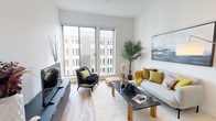 This well-appointed single-floor residence offers a cohesive blend of modern design and functional living. The open-concept living area is bathed in natural light, courtesy of expansive floor-to-ceiling sliding windows that frame views of the surrounding building exterior. Light hardwood flooring runs throughout the main living spaces, complementing the clean white walls and creating a bright, airy ambiance. A wall-mounted mini-split system ensures year-round climate comfort, while recessed lighting adds a polished touch.

The kitchen is a standout feature, showcasing rich walnut-toned cabinetry that extends to the ceiling for ample storage, paired with a sleek grey glass backsplash, white quartz countertops, and a full suite of stainless steel appliances including a range, microwave, and dishwasher.

The bathroom continues the warm, contemporary aesthetic with walnut cabinetry, an undermount sink, and white quartz surfaces. A stacked in-unit washer and dryer is conveniently integrated into the bathroom space, adding everyday practicality. The bedroom, visible in the layout, benefits from the same refined finishes and easy access to the main living corridor. Overall, the space delivers a clean, modern lifestyle with thoughtful details at every turn.