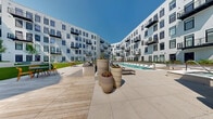Located at 1 Gibson Way in Revere, MA, this expansive outdoor amenity courtyard is nestled within a contemporary multi-story residential complex defined by crisp white horizontal siding, black-framed windows, and private balconies. The centerpiece of the space is a generously sized resort-style swimming pool with crystal-clear blue water, flanked by a row of sleek lounge chairs and complemented by poolside umbrellas for shade. A striking wood-beam pergola anchors one end of the pool deck, casting dramatic linear shadows across the light-toned stone pavers and adding an architectural focal point to the outdoor experience. Adjacent to the pool area, a dedicated lawn courtyard features lush green turf framed by raised planting beds filled with ornamental grasses, shrubs, and young trees, creating a tranquil garden setting. A separate outdoor lounge area offers modern seating arrangements on a combination of wood decking and large-format stone tile, ideal for casual gatherings. Floor-to-ceiling glass-walled corridors connect interior amenity spaces to the courtyard, maintaining a seamless indoor-outdoor flow. Large decorative planters with seasonal plantings add warmth and texture throughout, while the open sky above floods the entire courtyard with abundant natural light.