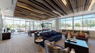 This contemporary amenity lounge is defined by its most dramatic architectural feature: a sculptural, undulating ceiling composed of layered wood-toned fins that ripple across the entire space, creating a bold visual statement and a sense of dynamic movement overhead. The open, single-floor layout is generously proportioned, offering distinct yet interconnected zones designed for socializing and relaxation.

Floor-to-ceiling windows and glass doors wrap around a significant portion of the lounge, flooding the interior with abundant natural light and providing expansive views of the surrounding streetscape and greenery. The light hardwood-style flooring complements the warm tones of the ceiling installation and grounds the space with a cohesive material palette.

A central seating area features a large navy blue sofa paired with eclectic, patterned accent chairs, creating an inviting conversation hub. Nearby, a full-size pool table serves as both a functional game area and a design focal point. A built-in wet bar and kitchenette — appointed with granite countertops, dark cabinetry, stainless steel appliances, a beverage refrigerator, and a striking chevron-patterned stone backsplash — adds excellent functional amenity to the space. Bar-height seating and a wall-mounted display screen complete the entertainment offerings. Curated artwork, lush indoor plants, and carefully chosen decorative accessories elevate the overall ambiance throughout.
