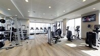 This fitness center offers a well-appointed workout environment designed to accommodate a variety of exercise routines. The space is flooded with natural light through expansive floor-to-ceiling windows that frame sweeping city skyline views, creating an energizing atmosphere for exercise. The ceiling features a clean, paneled design with recessed lighting that supplements the abundant natural light throughout the day.

The cardio area is well-stocked with a row of treadmills positioned directly in front of the large windows, allowing users to enjoy panoramic views during their workout. Alongside the treadmills, multiple elliptical machines and other cardio equipment are strategically arranged to maximize the use of space while maintaining comfortable spacing between machines. Each cardio unit appears to be equipped with individual display screens.

A dedicated strength-training area includes a dumbbell rack stocked with a range of free weights, as well as functional cable and weight machines. The flooring transitions between carpet and rubberized fitness mats, clearly delineating workout zones. The overall design is contemporary and clean, with a neutral color palette of whites and greys contributing to a professional and motivating fitness environment.