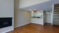 This two-story space showcases an open and airy design that promotes a seamless flow between the living and kitchen areas. The main living area features warm-toned hardwood flooring that extends throughout, complementing the neutral beige walls and crisp white baseboards. Elevated ceilings contribute to a notably spacious feel, enhancing the sense of openness throughout the main level.

A ceiling fan with integrated lighting is centrally positioned above the living area, providing both functionality and a focal point for the room. The kitchen is defined by a raised breakfast bar peninsula that serves as a natural divider between the two areas while maintaining visual connectivity. White cabinetry lines the kitchen walls, paired with built-in appliances including a microwave and range. The kitchen also benefits from its own overhead lighting fixture.

A doorway visible from the main living space hints at additional rooms beyond, while a partial view of what appears to be a closet or storage area adds to the overall utility of the layout. The clean, neutral palette and consistent flooring create a cohesive and welcoming atmosphere throughout the main level of this two-story space.