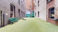 Located in the heart of Baltimore, MD (21201), this outdoor dog park is a thoughtfully designed urban retreat tucked within a courtyard formed by the towering brick facades of surrounding multi-story historic buildings. The open-air space is fully surfaced with lush artificial turf, providing a clean and comfortable area for dogs to roam and play year-round.

The park is enclosed by dark green chain-link fencing with a double-gated entry, ensuring safe access and containment for pets. A standout visual feature is a large, vivid mural painted across one of the brick walls, depicting a serene waterscape with sailboats, rolling green hills, and an open sky — adding a striking artistic element to the urban environment.

The space is furnished with green park benches positioned along the perimeter, offering seating for pet owners. Several large decorative boulders are placed throughout the turf, serving as both aesthetic accents and enrichment features for dogs. A waste station with bag dispensers and a trash receptacle are conveniently located near the entry, supporting cleanliness and responsible pet ownership.

The surrounding historic brick architecture rises several stories on all sides, creating a sheltered, enclosed atmosphere while still allowing open sky above. This urban dog park effectively combines functional amenities with creative design to serve both pets and their owners in a downtown Baltimore setting.
