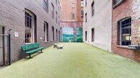 Located in the heart of Baltimore, MD (21201), this outdoor dog park is a thoughtfully designed urban retreat tucked within a courtyard formed by the towering brick facades of surrounding multi-story historic buildings. The open-air space is fully surfaced with lush artificial turf, providing a clean and comfortable area for dogs to roam and play year-round.

The park is enclosed by dark green chain-link fencing with a double-gated entry, ensuring safe access and containment for pets. A standout visual feature is a large, vivid mural painted across one of the brick walls, depicting a serene waterscape with sailboats, rolling green hills, and an open sky — adding a striking artistic element to the urban environment.

The space is furnished with green park benches positioned along the perimeter, offering seating for pet owners. Several large decorative boulders are placed throughout the turf, serving as both aesthetic accents and enrichment features for dogs. A waste station with bag dispensers and a trash receptacle are conveniently located near the entry, supporting cleanliness and responsible pet ownership.

The surrounding historic brick architecture rises several stories on all sides, creating a sheltered, enclosed atmosphere while still allowing open sky above. This urban dog park effectively combines functional amenities with creative design to serve both pets and their owners in a downtown Baltimore setting.