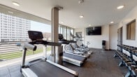 Located at 200 W Norwich Ave in Columbus, Ohio, this well-equipped fitness center offers a comprehensive workout environment designed to support a wide range of exercise routines. The space is thoughtfully laid out across a single floor, with distinct zones for cardio training, strength work, and free weights.

Along one side of the room, a row of cardio machines — including treadmills, ellipticals, and a stationary spin bike — lines the expansive floor-to-ceiling windows, allowing users to exercise with ample natural light and open views of the surrounding Columbus neighborhood. The large glazed panels give the space an airy, open feel that enhances the overall workout experience.

The strength training area features a Rogue power rack with a barbell and weight plates, along with multiple adjustable benches for varied lifting exercises. A full dumbbell rack with an organized range of free weights provides additional versatility. Floor-to-ceiling mirrors line one wall, making the space feel larger while allowing users to monitor their form.

Underfoot, dark rubber flooring covers the entire gym, providing both durability and safety during high-impact activities. Recessed lighting throughout ensures the space is well-lit at all times. A wall-mounted flat-screen TV and a standing water dispenser add practical amenities, completing this modern, fully outfitted fitness facility.