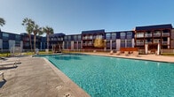 This multi-story residential complex showcases a striking exterior characterized by bold blue and white facade panels, arched balcony detailing, and large windows that lend the building a modern coastal aesthetic. The complex is arranged in a U-shaped configuration, creating an enclosed courtyard that hosts a generously sized swimming pool as its central amenity. The pool's clear turquoise water is surrounded by a broad concrete deck, providing ample space for lounging and recreation.

The outdoor amenity area is thoughtfully appointed with multiple lounge chairs positioned both in open sun and beneath a pergola-style shade structure, offering residents flexibility in how they enjoy the outdoor space. A dedicated outdoor kitchen and BBQ station, sheltered under a wooden pergola strung with ambient lighting, provides a functional and inviting gathering point for cooking and entertaining. Tall palm trees are planted throughout the courtyard, adding a tropical character to the setting and complementing the clear, sunny climate.

A manicured green lawn area is enclosed within a white perimeter fence, offering an additional recreational space adjacent to the pool deck. The overall outdoor environment is designed to encourage an active, social lifestyle, with each amenity zone flowing naturally into the next under open skies.