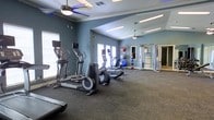 This fitness center offers a comprehensive workout environment designed to accommodate a variety of exercise routines. The space is finished with durable dark carpeting throughout, providing a stable and comfortable surface for both machine-based and floor exercises. The walls are painted in a cool blue-gray tone, contributing to a calm and focused atmosphere.

The cardio zone features multiple elliptical trainers and a stationary bike, arranged along the windowed wall to allow natural light to flow in during workouts. Large windows on two sides of the room enhance the sense of openness and provide ventilation alongside ceiling fans, which help maintain airflow throughout the space. The ceiling fans are equipped with integrated lighting, supplemented by recessed fluorescent fixtures overhead.

Along the far wall, a multi-functional cable and weight machine offers options for strength training. An assortment of exercise balls and additional accessories are neatly stored in a rack nearby. A doorway leads to an adjoining room, suggesting additional facilities may be available. Crown molding along the ceiling adds a polished architectural detail to the overall design.