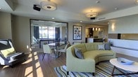 This contemporary single-floor amenity space is thoughtfully designed to foster social interaction and relaxation. The main lounge area centers around a curved, sage-green sectional sofa accented with decorative pillows, positioned atop a striped area rug that adds a layer of visual interest to the wood-style flooring. Recessed lighting and flush-mount drum pendant fixtures illuminate the space with a warm, inviting glow.

A dedicated dining area features a sleek rectangular table with upholstered chairs, ideal for casual gatherings or collaborative use. Adjacent to the lounge, a glass-partitioned billiards room provides a recreational retreat, enhanced by large windows that flood the area with natural light and offer open exterior views.

Toward the back of the space, a stylish bar counter with pendant lighting and dark cabinetry serves as both a functional and aesthetic focal point. Framed wall art and carefully selected décor contribute to the space's polished, modern character.

The open, flowing layout connects each zone seamlessly, creating a versatile environment suited for both quiet lounging and lively social events. The neutral color palette, punctuated by earthy tones and contemporary furnishings, gives the space a refined yet approachable ambiance throughout.