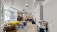 This well-appointed single-floor amenity space combines comfort and functionality in a thoughtfully designed layout. The main lounge area anchors the space with a curved white sofa, modern accent chairs, and a bold yellow modular ottoman arrangement set atop a graphic black-and-white area rug. Tray ceilings with crown molding and sleek black ceiling fans add architectural character overhead, while recessed lighting ensures a bright, welcoming atmosphere throughout.

Large windows along the exterior walls invite generous natural light and offer views of the surrounding greenery, enhancing the open feel of the space. A raised bar-height workstation with a corrugated metal base and dual computer monitors serves as a dedicated co-working area, accented by a vibrant Texas-themed art print above. Along one wall, a built-in banquette upholstered in navy blue provides additional seating alongside a row of round café tables and stools, ideal for casual gatherings or focused work.

Curated wall art, including abstract framed prints and a striking neon piece, adds a creative edge to the overall design. A cluster of organic-shaped decorative mirrors further enlivens the lounge. A separate conference room features a large oval wood table, colorful modern chairs, a glass whiteboard, and windows overlooking lush outdoor trees, making it a practical space for meetings or private study.