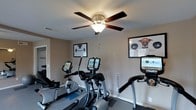 This dedicated fitness space is thoughtfully designed to support an active lifestyle within the comfort of home. The room is outfitted with multiple cardio machines, including a recumbent bike, an elliptical trainer, and a treadmill, each equipped with its own digital display screen for tracking workout metrics. Neutral-toned walls create a clean, focused environment, while a five-blade ceiling fan with a central light fixture ensures steady airflow and comfortable illumination throughout exercise sessions. A large flat-screen monitor is mounted on one wall, providing an excellent option for streaming workout content or entertainment. The space is further personalized with framed wall art featuring a playful dog weightlifting theme, adding a lighthearted character to the room. The flooring is finished in a dark tone suitable for high-traffic fitness use. A full-length mirror along one wall enhances the sense of space and allows for form-checking during workouts. Natural light filters in through a nearby window, keeping the atmosphere bright and inviting. Overall, this fitness room offers a well-organized, functional, and motivating environment for daily exercise routines.